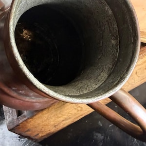 May include: A copper pitcher with a wide, circular opening and a metallic handle. The interior of the vessel features a textured, silver-toned lining that contrasts with the smooth, warm-toned copper exterior. The pitcher has a cylindrical body.