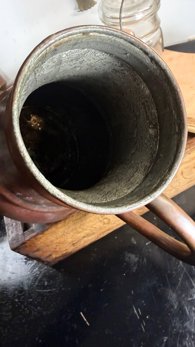 May include: A copper pitcher with a wide, circular opening and a metallic handle. The interior of the vessel features a textured, silver-toned lining that contrasts with the smooth, warm-toned copper exterior. The pitcher has a cylindrical body.