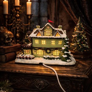 May include: A lighted Christmas village house with a green exterior, snow-covered roof, and a small red flag. The house has multiple lit windows and sits on a wooden table. A small Christmas tree and lit candles are in the background.