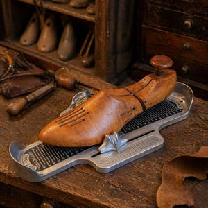May include: A wooden shoe last with a knob is placed on a metal shoe measuring device. The shoe last is light brown with dark brown markings. The measuring device has black and white markings and is silver. The background includes wooden shelves and drawers.