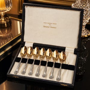 Puede incluir: Un juego de siete cucharas doradas con mangos de plata ornamentados, dispuestas en una caja de presentación negra. La tapa interior muestra el texto: Made in Sheffield, England for Bergdorf Goodman. Las cucharas descansan sobre un forro de tela crema.