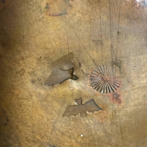 May include: A weathered, circular copper surface featuring a metallic patina with visible scratches and oxidation. The surface is embossed with three distinct stamped designs: a small sunburst pattern, a bat silhouette, and a fish shape.