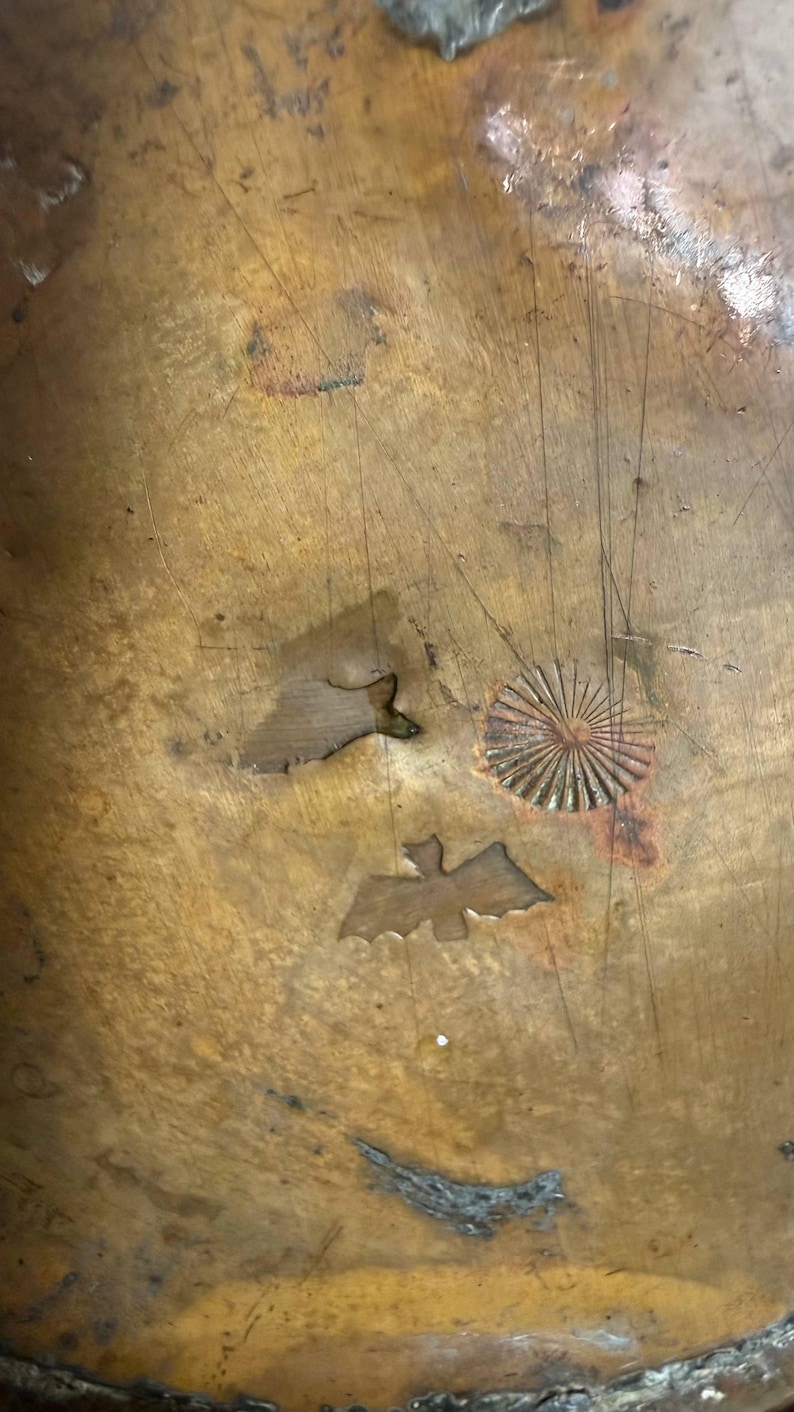 May include: A weathered, circular copper surface featuring a metallic patina with visible scratches and oxidation. The surface is embossed with three distinct stamped designs: a small sunburst pattern, a bat silhouette, and a fish shape.