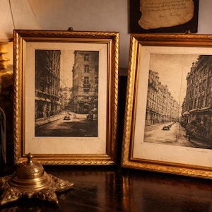 May include: Two framed black and white prints of a European street scene with gold ornate frames. A lit candle sits in a wine bottle, and a decorative bell is on the table. The prints are vintage-style.