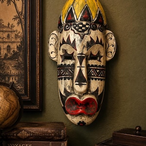 Puede incluir: Una máscara de pared de madera tallada a mano con un acabado crema desgastado y patrones geométricos pintados en negro, rojo y amarillo. La máscara tiene una parte superior redondeada, orejas prominentes, ojos rectangulares y labios rojos.