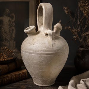 May include: A cream-colored, antique-style ceramic jug with a large handle and two spouts. The jug has a textured surface and a rounded body. It is placed on a dark wooden surface with other vintage items in the background.
