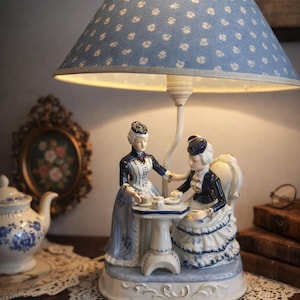 Könnte beinhalten: Dekorative Tischlampe mit blauem Lampenschirm mit weißen Blumenmustern. Der Sockel zeigt zwei Porzellanfiguren in viktorianischen Kleidern an einem Tisch. Eine Teekanne und ein gerahmtes Kunstwerk sind sichtbar.