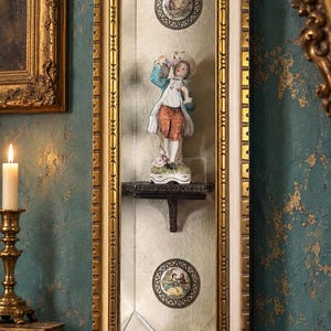 May include: An elongated octagonal wall mirror with a gold and cream frame, featuring a central shelf holding a porcelain figurine of a person in a blue coat and orange breeches, flanked by two circular floral medallions set against a silvered glass.