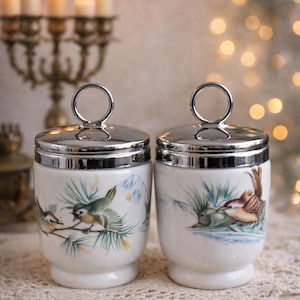 May include: Two white ceramic egg cups with silver lids and handles. Each cup features a hand-painted design of birds perched on branches with green foliage. The background includes a mountain scene.