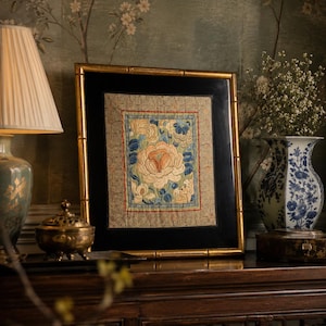May include: A square framed textile art piece featuring intricate embroidery of a central peach-colored peony surrounded by blue floral motifs. The embroidery is set against a beige patterned background, enclosed by a black mat and a gold bamboo-style frame.