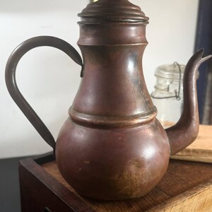 May include: A copper teapot with a rounded base, a tapered neck, and a matching lid with a small knob handle. The teapot features a curved spout and a thin, arched handle attached to the side. The metal surface shows a weathered, dark patina.
