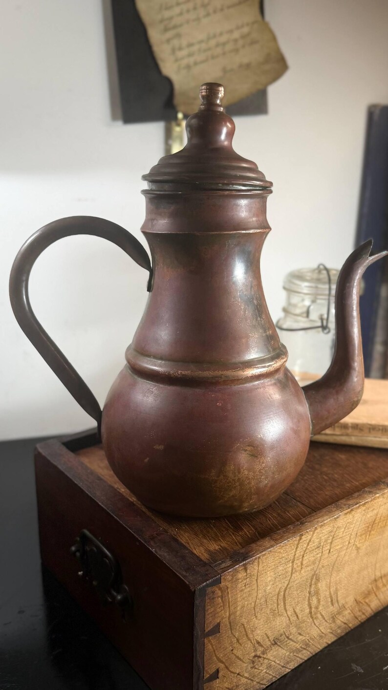 May include: A copper teapot with a rounded base, a tapered neck, and a matching lid with a small knob handle. The teapot features a curved spout and a thin, arched handle attached to the side. The metal surface shows a weathered, dark patina.