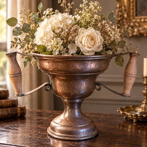 May include: A copper vessel with a wide, flared rim and a pedestal base, featuring two vertical wooden handles attached by curved metal brackets. The vessel is filled with a bouquet of white roses, hydrangeas, and small green foliage.