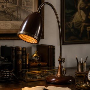 Puede incluir: Una lámpara de escritorio con una pantalla en forma de cúpula de color marrón oscuro y un brazo curvo de latón. La lámpara cuenta con una base circular y una articulación ajustable con un pomo en forma de engranaje. El interior tiene una rejilla.