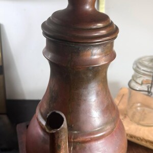 May include: A tall, weathered copper coffee pot with a rounded body, a narrow neck, and a matching lid featuring a small knob. The metal surface displays a dark, oxidized patina with visible signs of age, including varied brown and reddish tones.