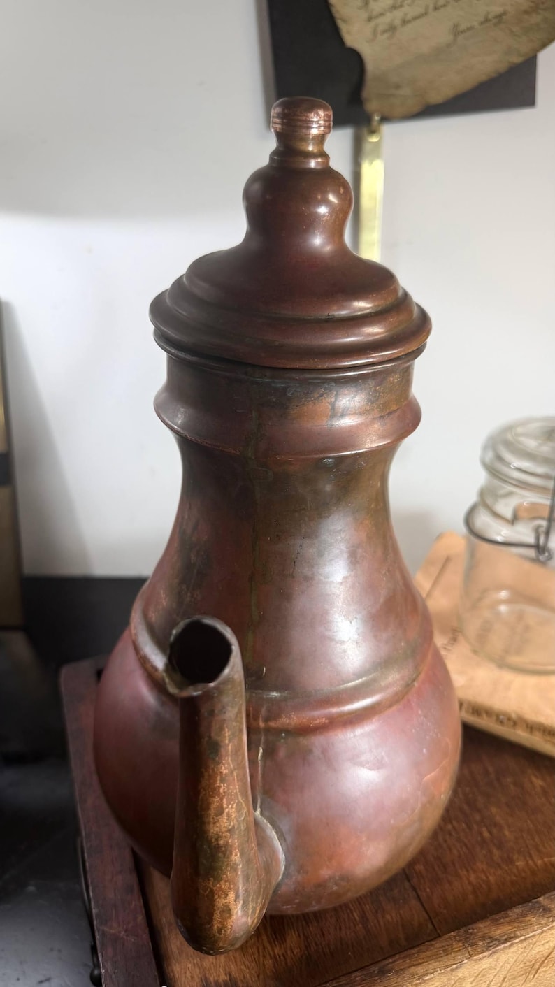 May include: A tall, weathered copper coffee pot with a rounded body, a narrow neck, and a matching lid featuring a small knob. The metal surface displays a dark, oxidized patina with visible signs of age, including varied brown and reddish tones.