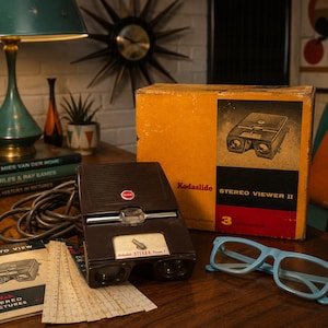 Op de afbeelding: Een bruine Kodak Stereo Viewer II met bijbehorende geel-zwarte kartonnen doos, een gedrukte handleiding getiteld How to View en een lichtblauwe plastic bril. Het apparaat is voorzien van een rood Kodak-logo en een zilveren metalen hendel.
