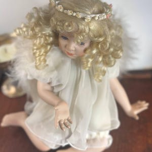 May include: A porcelain angel doll with blonde, curly hair and a floral crown. The doll wears a white dress with feather accents and is posed kneeling on a wooden surface. The doll's face has delicate features and blue eyes.