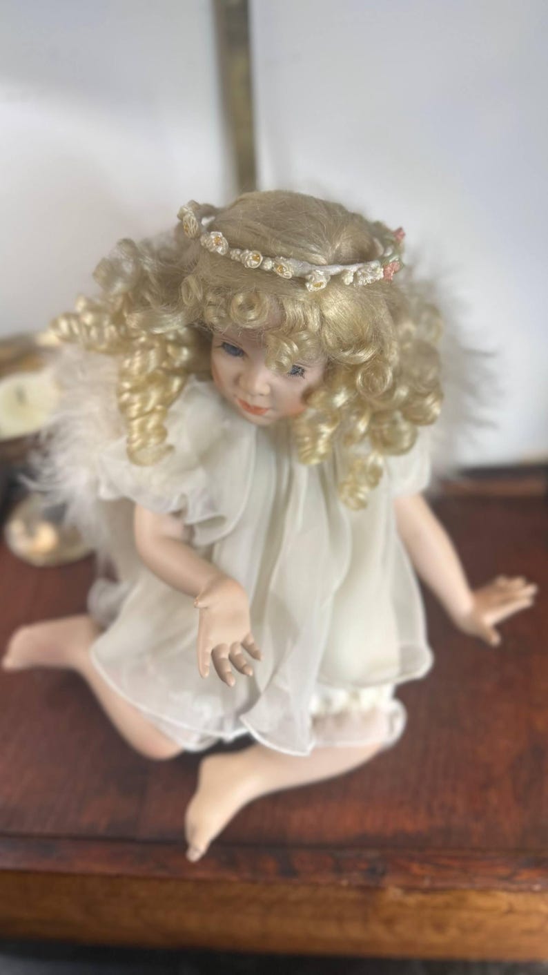 May include: A porcelain angel doll with blonde, curly hair and a floral crown. The doll wears a white dress with feather accents and is posed kneeling on a wooden surface. The doll's face has delicate features and blue eyes.