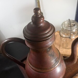 May include: A copper teapot with a rounded body, a curved spout, and a matching lid featuring a small knob handle. The metal surface displays a warm, weathered patina with dark brown and reddish-orange tones throughout the exterior finish.