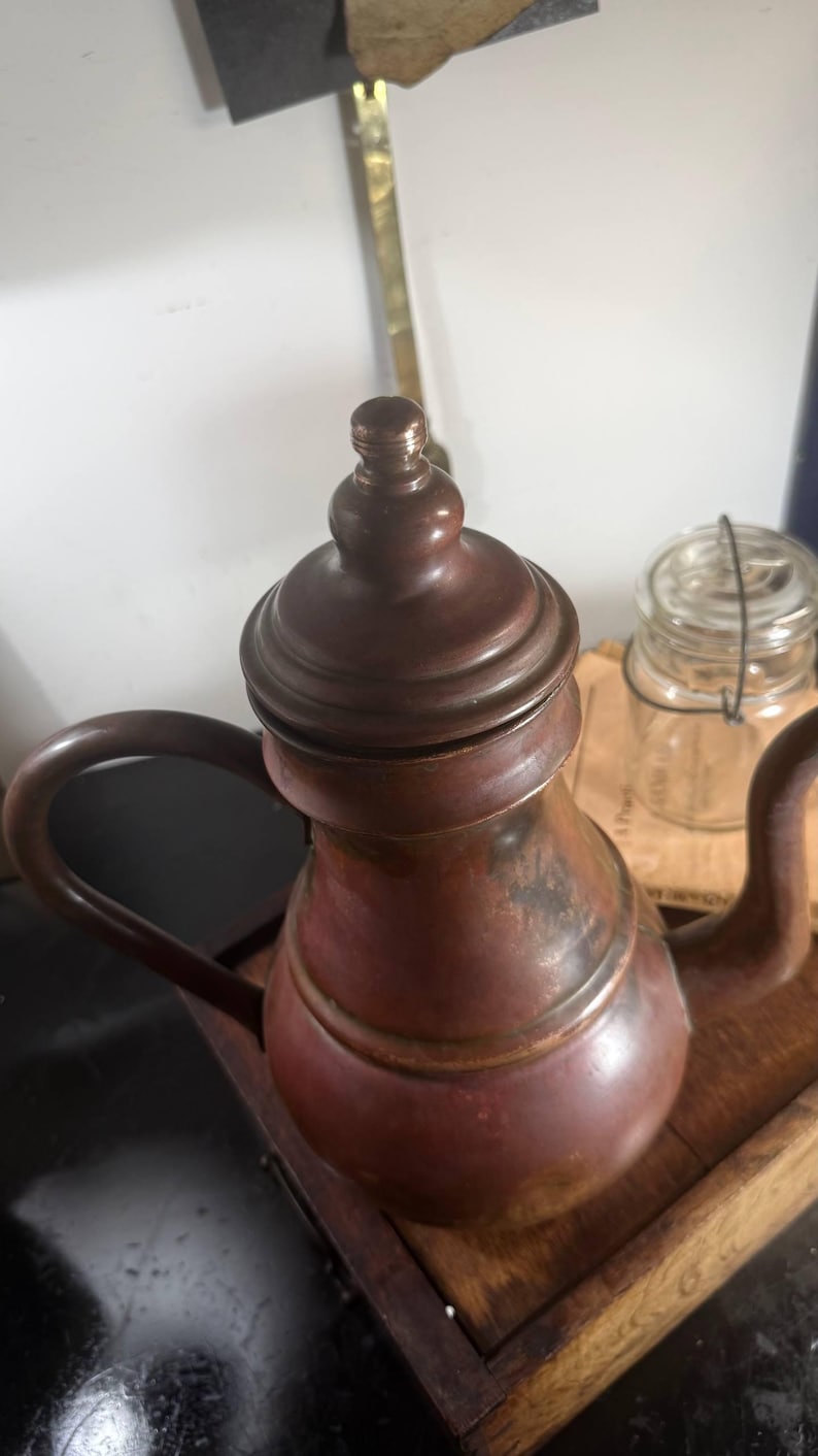 May include: A copper teapot with a rounded body, a curved spout, and a matching lid featuring a small knob handle. The metal surface displays a warm, weathered patina with dark brown and reddish-orange tones throughout the exterior finish.