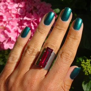 May include: A hand wearing a silver ring with a large red rectangular stone. The hand is painted with dark green nail polish.