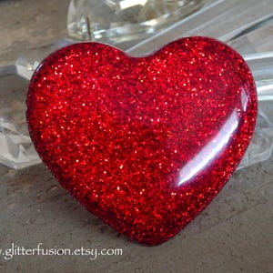 May include: A red glitter heart, reflecting light, with a glossy finish. The heart shape is the central focus, with a clear, faceted object in the background. The website address www.glitterfusion.etsy.com is visible.