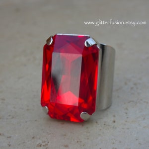 May include: A large, rectangular red gemstone ring with a silver-colored band. The gemstone is faceted, catching the light and displaying a vibrant red hue. The ring's design is simple yet striking, making it a bold statement piece.