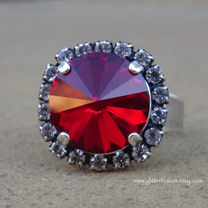 May include: A silver ring with a large, red, faceted crystal surrounded by a halo of smaller, clear crystals.