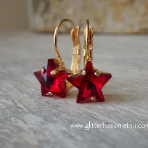 May include: A pair of gold-colored leverback earrings with star-shaped, faceted, red crystal pendants. The earrings are displayed on a light gray surface. The website address www.glitterfusion.etsy.com is visible in the image.