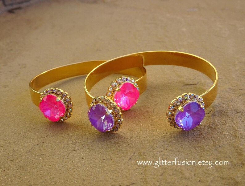 May include: Two gold bangle bracelets with jeweled accents. The bracelets feature a large pink rhinestone and a large purple rhinestone.