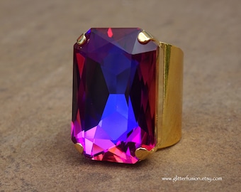 May include: A large, rectangular ring with a gold band and a vibrant, multi-colored gemstone. The gemstone transitions from red to purple to blue. The ring is a statement piece, perfect for adding a pop of color to any outfit.