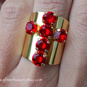 Op de afbeelding: Een goudkleurige ring met een kruisontwerp, bezet met helderrode edelstenen. De ring heeft een brede band en de edelstenen zijn rond. Het websiteadres www.glitterfusion.etsy.com is zichtbaar.