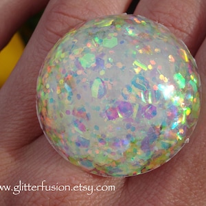 Può includere: Un oggetto sferico trasparente pieno di glitter iridescenti nei toni del verde, blu, viola e giallo. L'oggetto è tenuto in mano. L'indirizzo del sito web www.glitterfusion.etsy.com è visibile in basso.