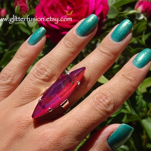 May include: A large, statement ring with a vibrant, elongated, faceted crystal in shades of pink and purple. The crystal is set in a silver-toned metal band. The ring is displayed on a hand with teal nail polish. The website address www.glitterfusion.etsy.com is visible.