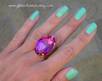 May include: A gold ring with a large, oval-shaped, fuchsia-colored rhinestone. The ring is worn on the middle finger of a hand with light green nail polish.