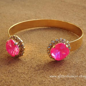 May include: A gold bangle bracelet with two large pink rhinestones set in clear crystal accents. The bracelet is open and has a simple design.