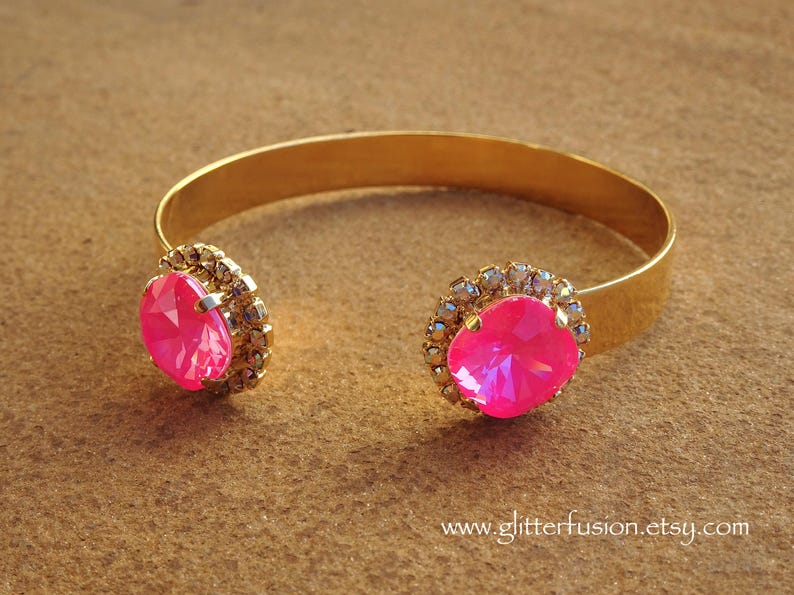 May include: A gold bangle bracelet with two large pink rhinestones set in clear crystal accents. The bracelet is open and has a simple design.