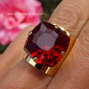 May include: A large, square-cut, ruby-red gemstone ring with a gold band. The ring features a bold, faceted stone held in place by four gold prongs. The website address www.glitterfusion.etsy.com is visible in the background.