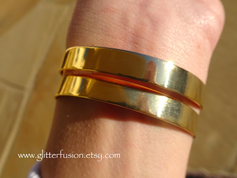 May include: Two gold-toned metal bangles stacked on a person's wrist. The bangles are smooth and shiny.