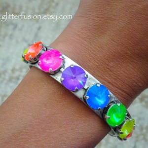 May include: A silver-toned bracelet with a row of large, colorful, round gemstones. The gemstones are in shades of neon orange, pink, purple, blue, and green. The bracelet is worn on a wrist. The website address "www.glitterfusion.etsy.com" is visible.