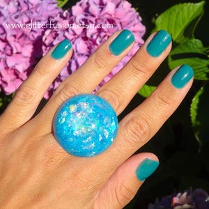 May include: A large, round, blue ring with iridescent glitter embedded within. The ring is set against a background of green foliage and pink flowers. The nails are painted a teal color. The website address www.glitterfusion.etsy.com is visible.