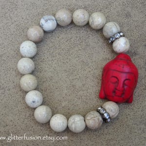 May include: A white stone beaded bracelet with a red Buddha charm. The bracelet has silver accents and is approximately 7 inches in length.