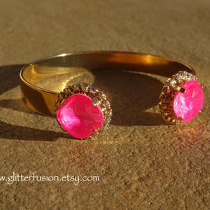 May include: A gold bangle bracelet with two large pink rhinestone accents. The rhinestones are surrounded by smaller clear rhinestones.