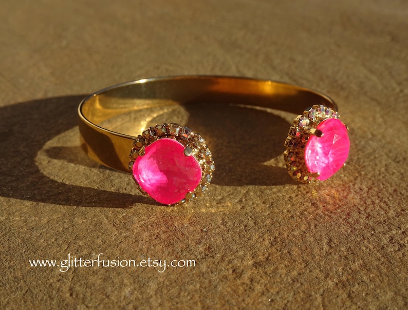 May include: A gold bangle bracelet with two large pink rhinestone accents. The rhinestones are surrounded by smaller clear rhinestones.