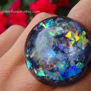 May include: A large, round ring with a dark base and embedded iridescent, colorful fragments. The ring's surface reflects light with hues of blue, green, and yellow. The website address "www.glitterfusion.etsy.com" is visible.