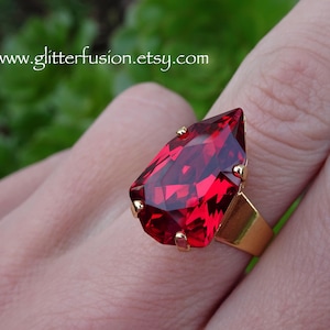 May include: A gold-toned ring featuring a large, faceted, teardrop-shaped red gemstone. The ring's band is a simple, rectangular design. The website address www.glitterfusion.etsy.com is visible in the background.
