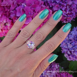 May include: A hand wearing a large, iridescent gemstone ring with a silver band. The nails are painted a shimmering teal color. The background features vibrant pink and purple flowers.