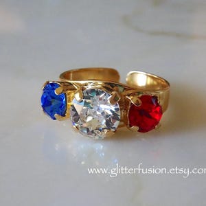 Può includere: Un anello regolabile color oro con tre pietre rotonde sfaccettate incastonate in griffe. La pietra centrale è trasparente, affiancata da una pietra blu a sinistra e una rossa a destra. Il testo www.glitterfusion.etsy.com è visibile in basso.