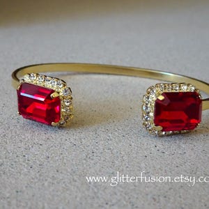 May include: Gold-toned cuff bracelet featuring two large, square, ruby-red gemstones. Each gemstone is surrounded by a halo of small, clear crystals. The bracelet has a simple, elegant design, perfect for adding a touch of glamour.